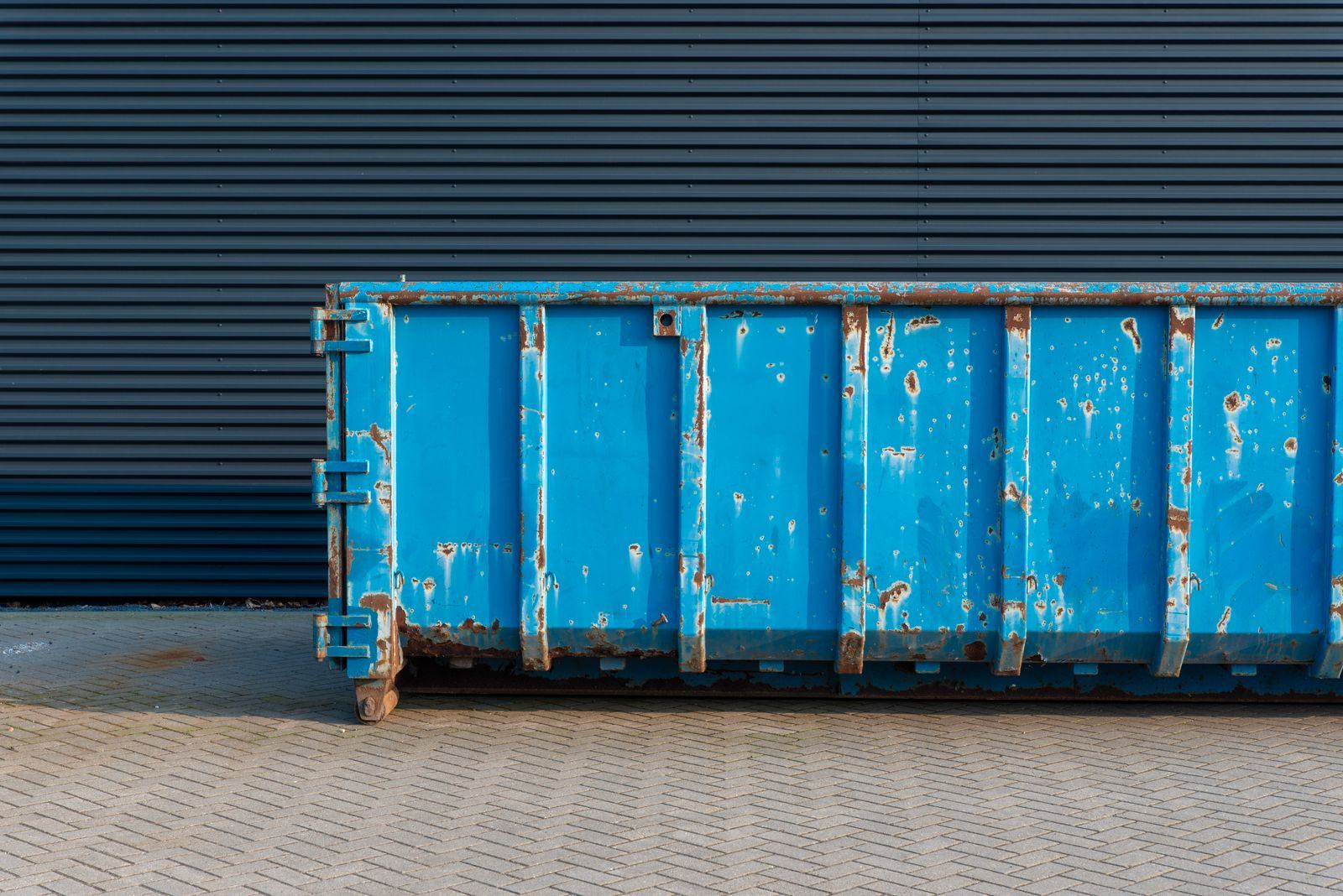 Weathered blue open-top roll-off container parked on a paved lot against a corrugated metal wall